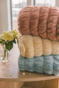 Stack of textured towels in pink, beige, and blue on a wooden surface with flowers in the background.