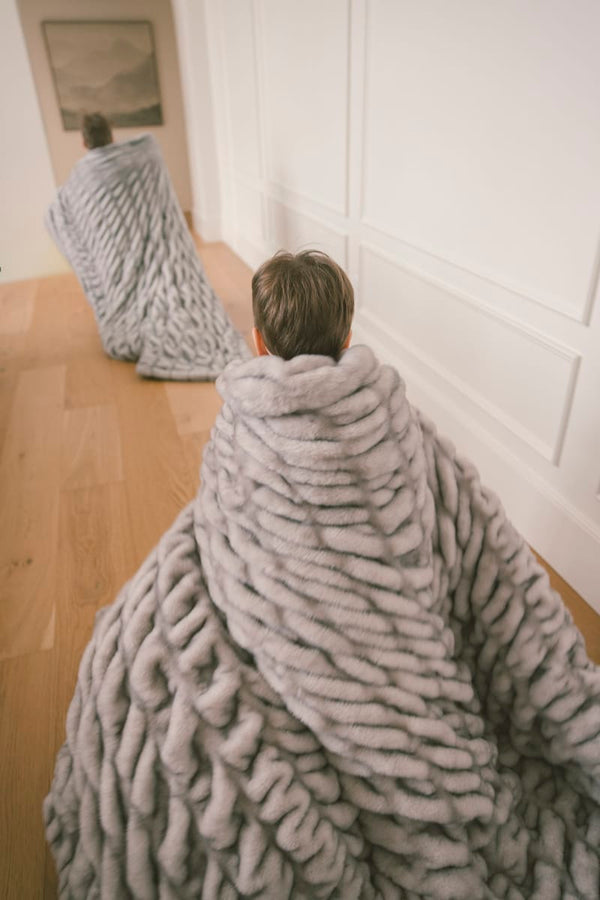 Two individuals wrapped in the cozy Hugs Chai Blanket, showcasing its soft gray and tan tones in a stylish indoor setting.