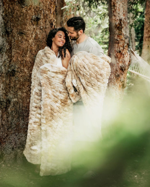 A couple embracing and smiling while wrapped in a large, fluffy Hugs Caramel Cotton Candy Minky Couture faux-fur blanket in a forest setting with large tree trunks behind them.