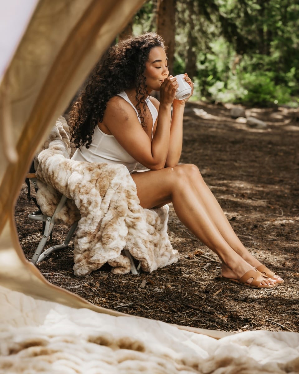 A woman with curly dark hair sits barefoot on a folding chair inside a tent, holding a white mug. A Hugs Cotton Candy Caramel blanket is draped over the chair, and a forest setting is visible in the background.
