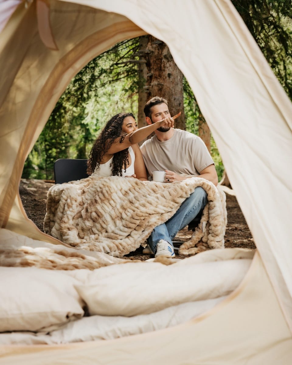 A couple relaxes inside a beige tent at a forest campsite, wrapped in a chunky k Hugs Cotton Candy Caramel blanket. The man wears a light gray t-shirt and blue jeans, and the woman wears a white top.