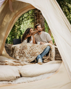 A couple relaxes inside a beige tent at a forest campsite, wrapped in a chunky k Hugs Cotton Candy Caramel blanket. The man wears a light gray t-shirt and blue jeans, and the woman wears a white top.