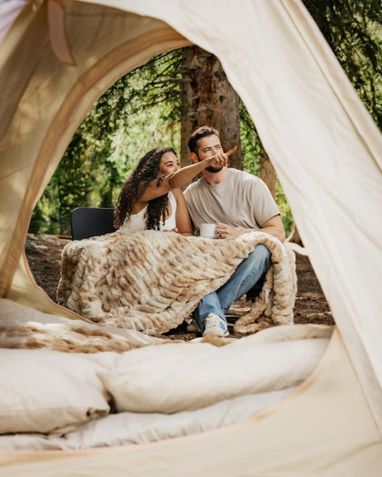 A couple relaxes inside a beige tent at a forest campsite, wrapped in a chunky k Hugs Cotton Candy Caramel blanket. The man wears a light gray t-shirt and blue jeans, and the woman wears a white top.
