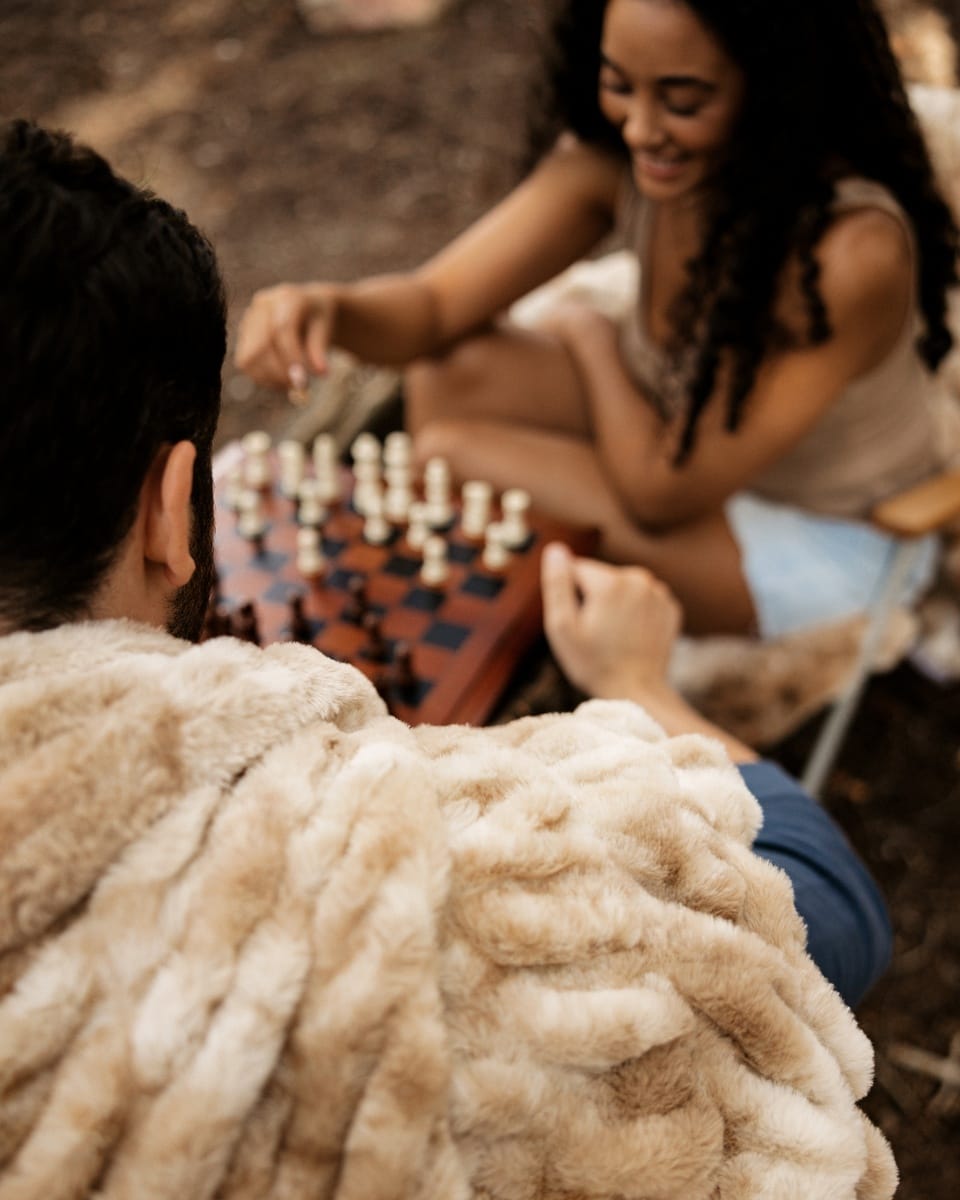 A cozy lifestyle scene of a couple playing chess outdoors. The person in the foreground is wearing a thick, fluffy Minky Couture Hugs Caramel blanket, while the person in the background is smiling and looking down at the chessboard.