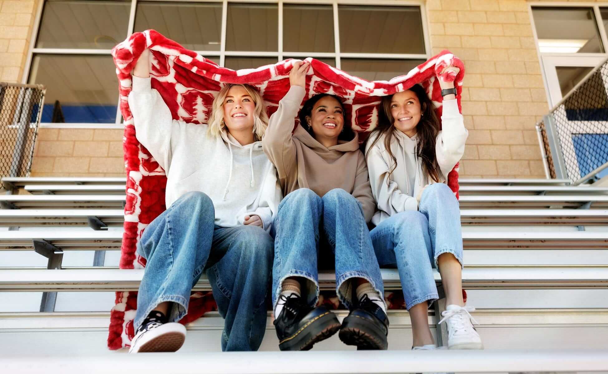 Three friends sitting on bleachers under a Hugs Stanford Cardinals Blanket, showcasing school spirit and comfort.