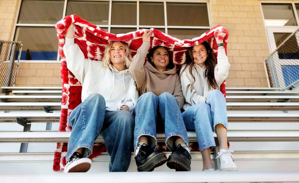 Three friends sitting on bleachers under a Hugs Stanford Cardinals Blanket, showcasing school spirit and comfort.