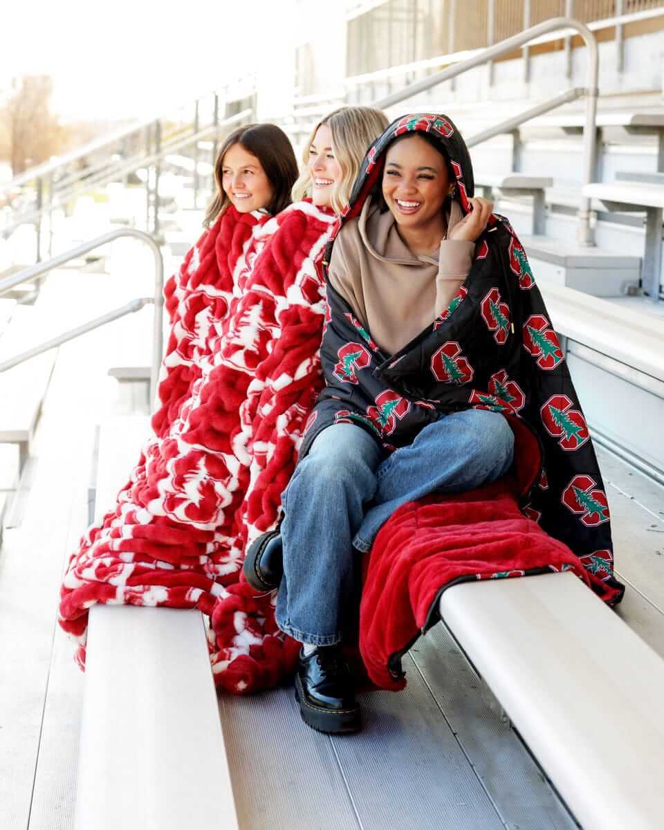 Three women wrapped in the Stanford Cardinals Hooded Stadium Blanket on stadium seats.