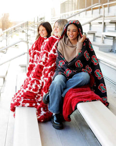 Three women wrapped in the Stanford Cardinals Hooded Stadium Blanket on stadium seats.