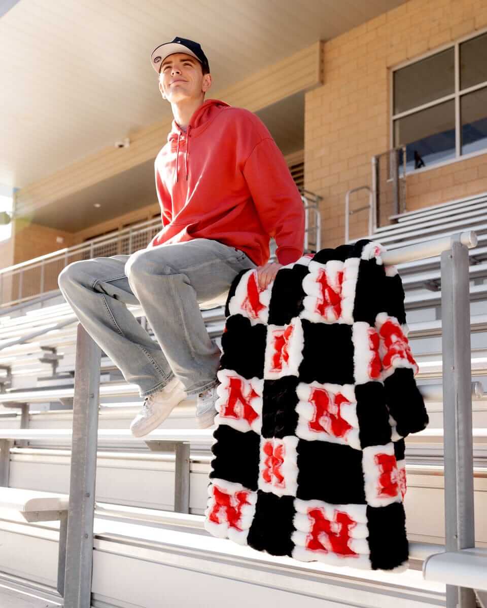 Nebraska Huskers Blanket draped over a seated fan, showcasing its black and white check pattern with red N logos.