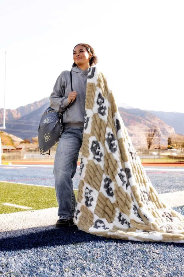 Woman on a football field draped in a Colorado Buffaloes Blanket - Hugs, showcasing its gold and cream check design.