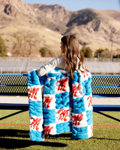 Hugs Ole Miss Rebels Blanket showcased outdoors with a girl wrapping it around, featuring a blue and white checkered design.