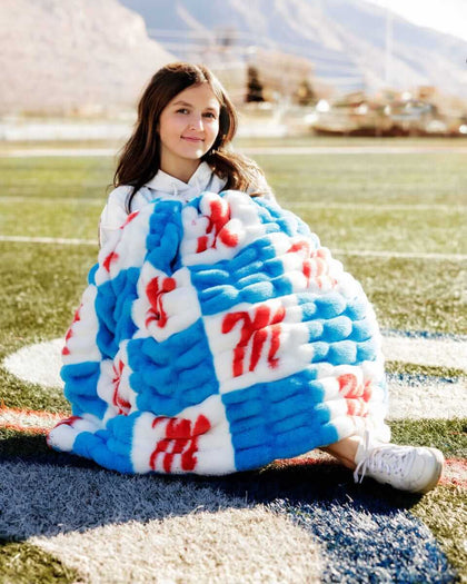 Girl wrapped in Ole Miss Rebels Blanket sitting on a sports field, showcasing its colorful blue and white checkered design.
