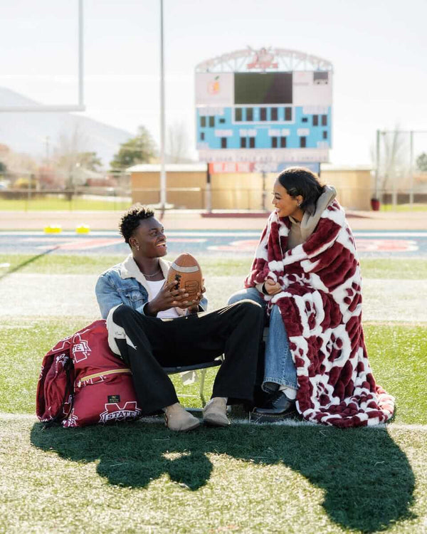 Couple on football field wrapped in Mississippi Hugs State Bulldogs Blanket, showcasing team pride and cozy comfort.