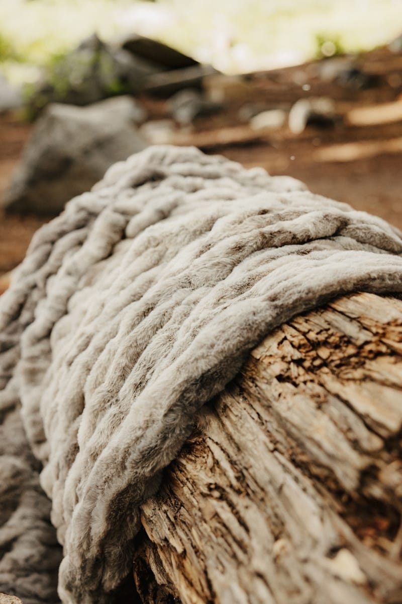 A chunky Hugs Cotton Candy Licorice gray blanket in a light gray color draped over a weathered log outdoors.