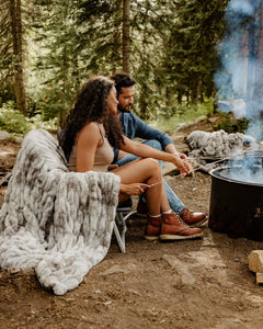 A couple sits by a campfire in a forest, roasting marshmallows on sticks. A chunky light gray Hugs Cotton Candy Licorice blanket is draped over a folding chair beside them, with trees and smoke in the background.