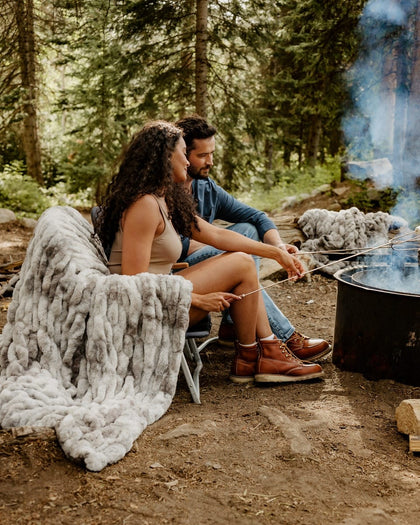 A couple sits by a campfire in a forest, roasting marshmallows on sticks. A chunky light gray Hugs Cotton Candy Licorice blanket is draped over a folding chair beside them, with trees and smoke in the background.