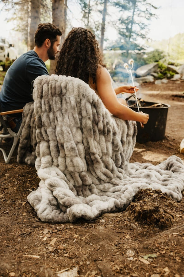 A person wrapped in a chunky, oversized Hugs Cotton Candy Licorice blanket sits outdoors at a campfire with another person beside them, holding a roasting stick. The setting is a wooded outdoor campsite with dirt ground, trees, and a black cooking pot in the background.