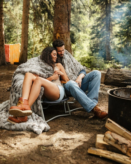 A couple relaxes in a camping chair wrapped in a Hugs Cotton Candy Licorice blanket outdoors in a forest setting, with a campfire and stacked logs nearby.