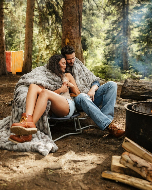 A couple relaxes in a camping chair wrapped in a Hugs Cotton Candy Licorice blanket outdoors in a forest setting, with a campfire and stacked logs nearby.