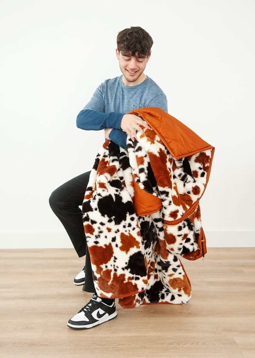 A person holding the Everywhere Blanket Brown Cow, showcasing its cow print and warm camel brown lining.