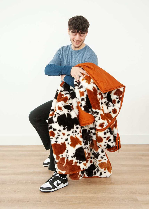 A person holding the Everywhere Blanket Brown Cow, showcasing its cow print and warm camel brown lining.