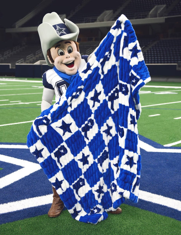 A Dallas Cowboys mascot stands on an indoor football field holding up a large blue and white checkered blanket with star and cowboy boot pattern.