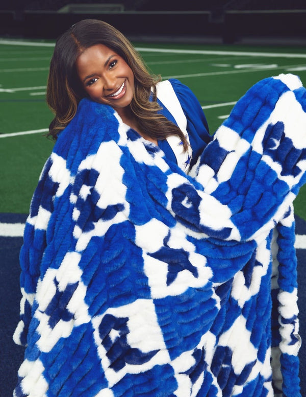 A smiling woman wrapped in a plush, oversized throw blanket featuring a bold royal blue and white pattern, posed on a green football field.