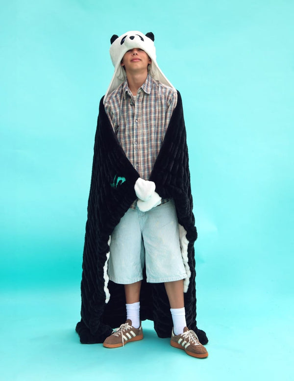 A person standing against a bright turquoise studio background wearing a plush Dude Perfect panda hooded blanket draped over their shoulders, a plaid button-up shirt, light denim shorts, white socks, and brown sneakers.