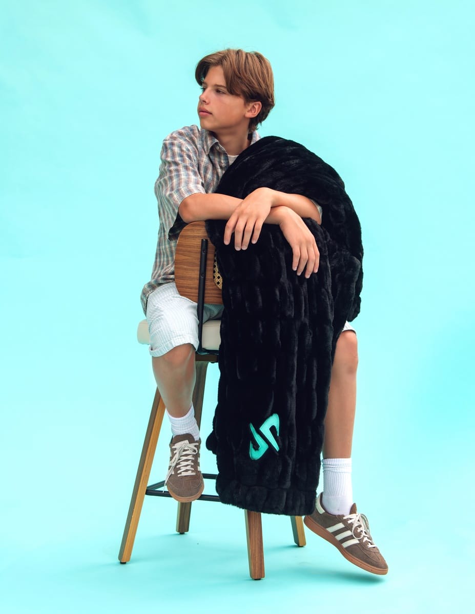 A person sitting on a wooden stool against a light blue studio background, wrapped in a black hugs Minky Couture and Dude Perfect blanket with a turquoise Dude Perfect logo. The person wears a plaid shirt, white shorts, socks, and brown sneakers.