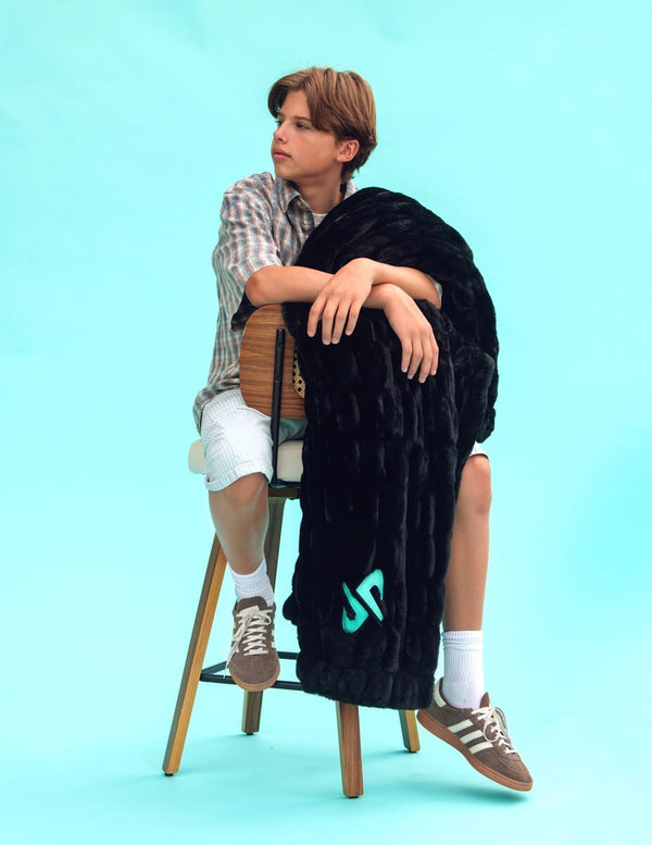 A person sitting on a wooden stool against a light blue studio background, wrapped in a black hugs Minky Couture and Dude Perfect blanket with a turquoise Dude Perfect logo. The person wears a plaid shirt, white shorts, socks, and brown sneakers.