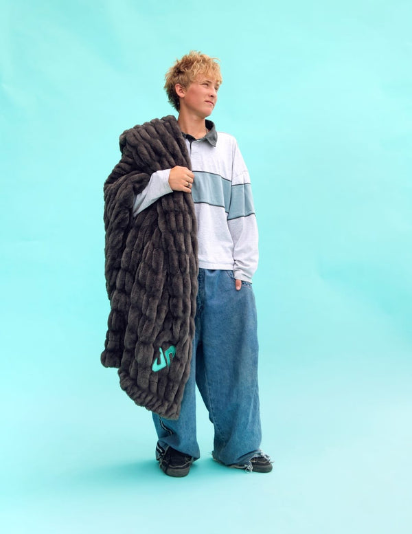 A model stands against a solid light blue studio background, holding a large, charcoal Minky Couture Dude Perfect Hugs blanket. The blanket features a small Dude Perfect logo near the bottom.