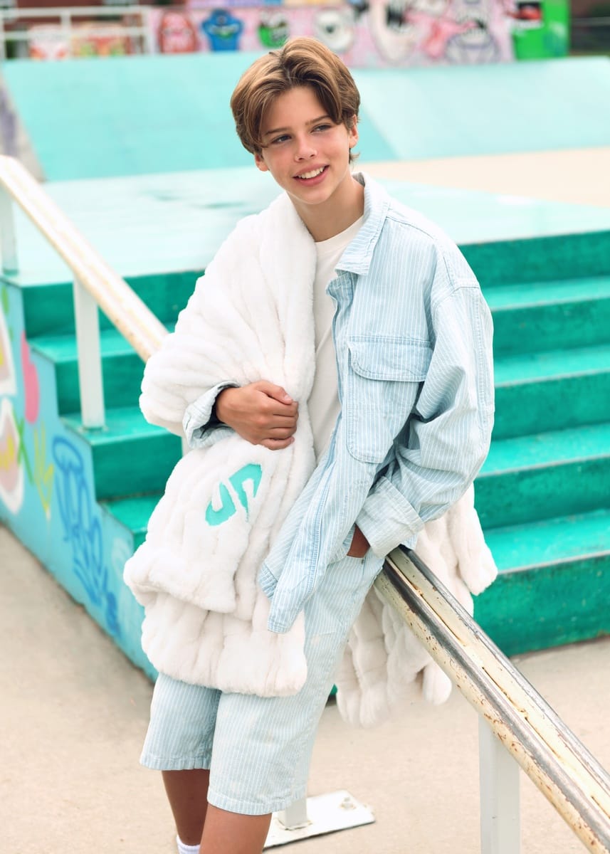 A young person standing outdoors in front of turquoise skatepark stairs, wearing a light blue pinstripe overshirt, matching shorts, and a cream Minky Couture Dude Perfect Hugs Cream blanket draped over their shoulders.