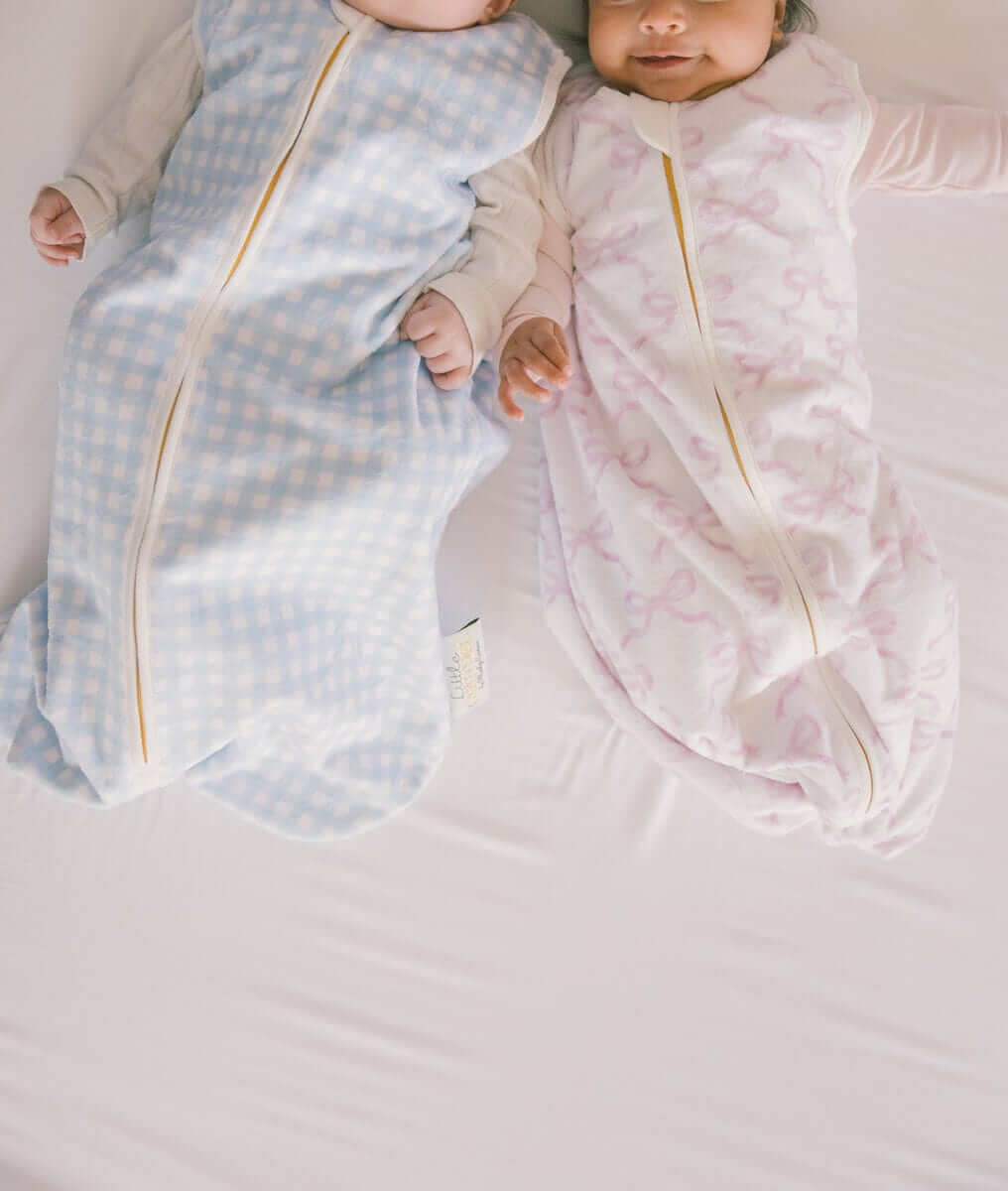 Two infants wearing Minky Couture Wearable Blankets and a pink version, on a white crib sheet.