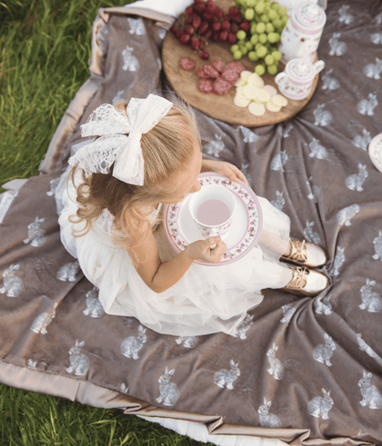 A child in a white dress enjoying tea on the Bunny Hop Milk Chocolate Blanket featuring playful bunny prints.