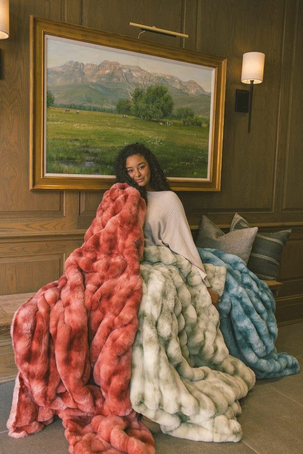 Young woman sitting on a wooden bench under a picture frame while wrapped up in three blanket. One blanket is red, one is light green, and the other is an ocean blue color.