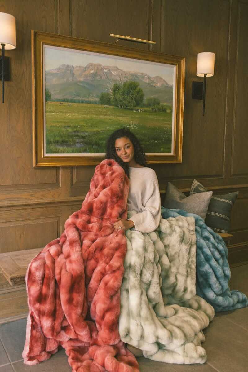 Woman sitting on a wooden bench wrapped up in three Extreme Ultra Cloud weighted blankets in red, light green and ocean blue color.