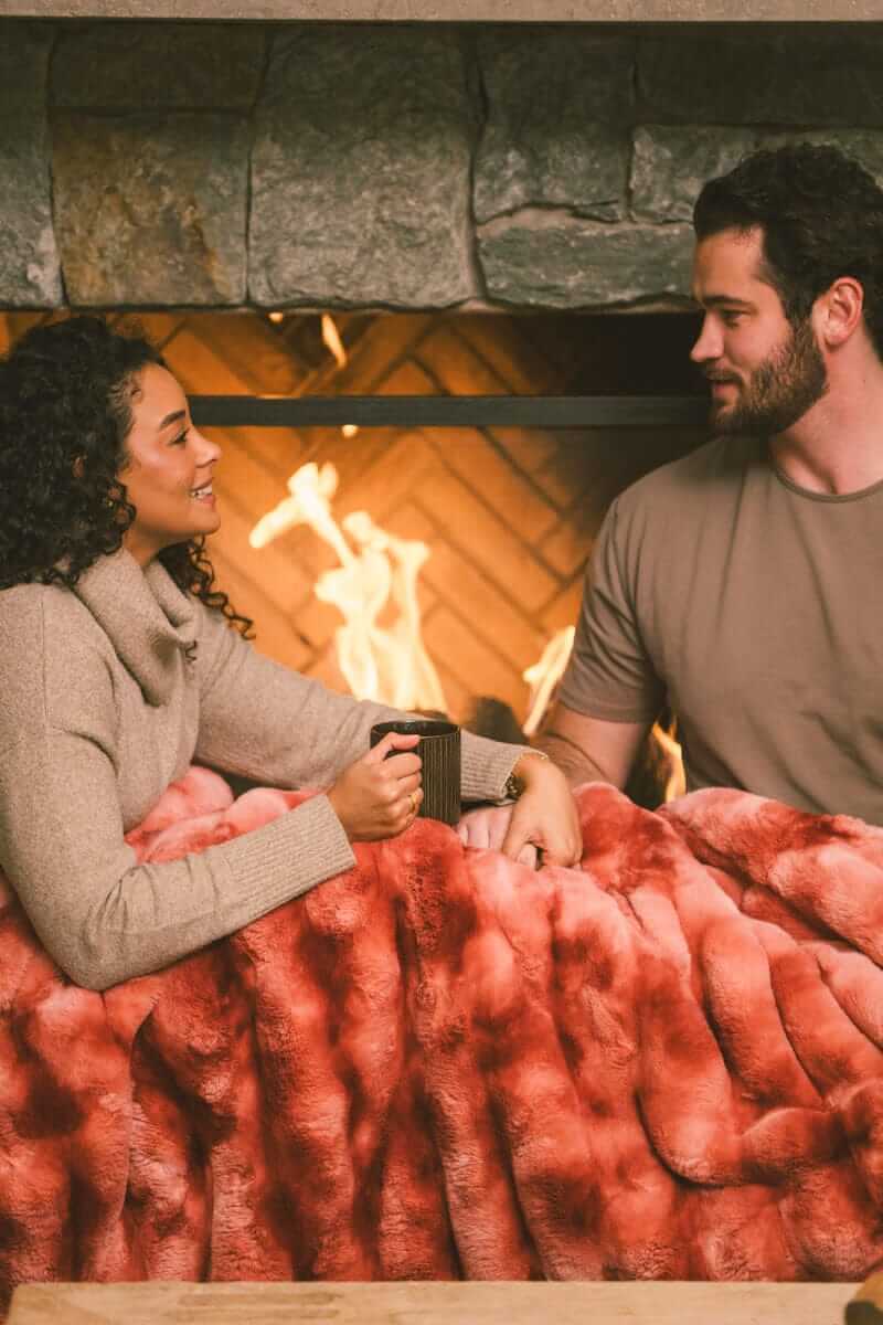 Couple sitting next to a fireplace while a Extreme Ultra Cloud Red Pear weighted blanket is draped over their laps.