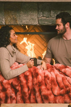 Couple sitting next to a fireplace while a Extreme Ultra Cloud Red Pear weighted blanket is draped over their laps.