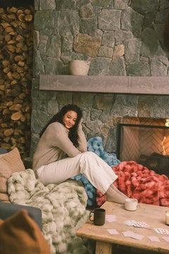 A cozy living room scene featuring a woman sitting with the Extreme Ultra Cloud Winter Sage Blanket, enjoying warmth by a fireplace.