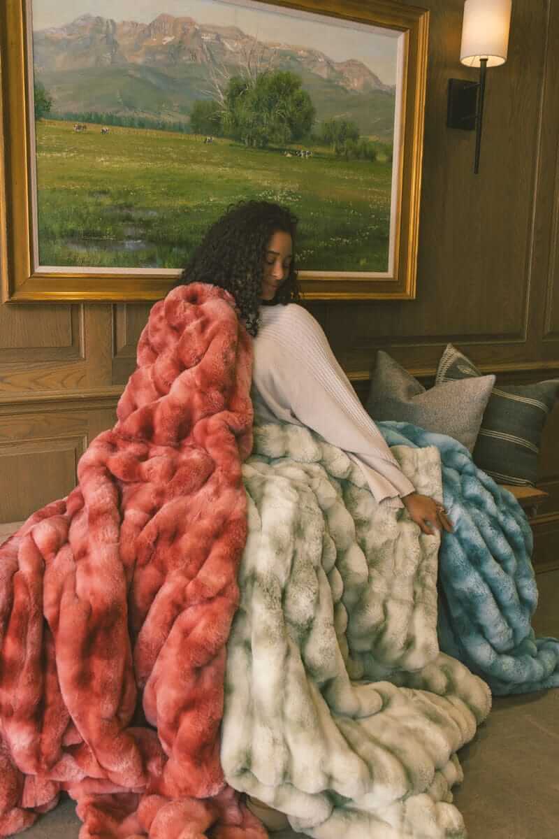 Woman sitting on a wooden bench wrapped up in three Extreme Ultra Cloud weighted blankets in red, light green and ocean blue color.