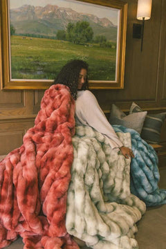 Woman sitting on a wooden bench wrapped up in three Extreme Ultra Cloud weighted blankets in red, light green and ocean blue color.