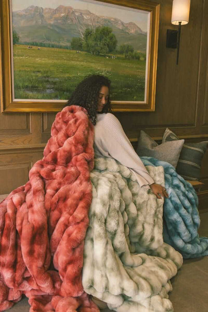 Woman sitting on a wooden bench wrapped up in three Extreme Ultra Cloud weighted blankets in red, light green and ocean blue color.