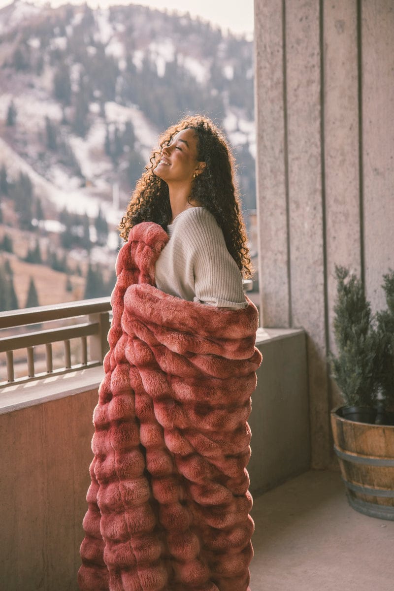 Young woman wrapped up in a two toned red blanket while standing on a balcony enjoying the view of the snowy mountains.