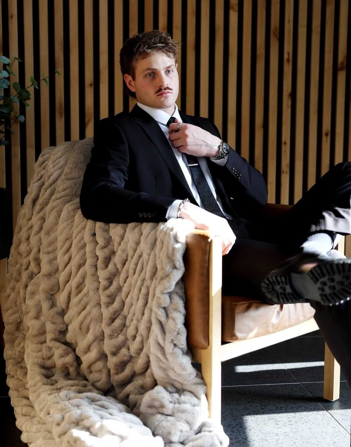 A man in a suit sitting on a chair, draped with a Biggest Hugs Tranquil Dove Blanket, showcasing its luxurious texture.