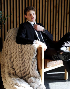 A man in a suit sitting on a chair, draped with a Biggest Hugs Tranquil Dove Blanket, showcasing its luxurious texture.