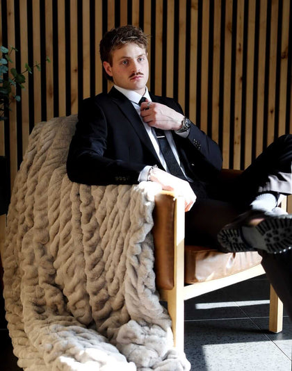 A man in a suit sitting on a chair, draped with a Biggest Hugs Tranquil Dove Blanket, showcasing its luxurious texture.