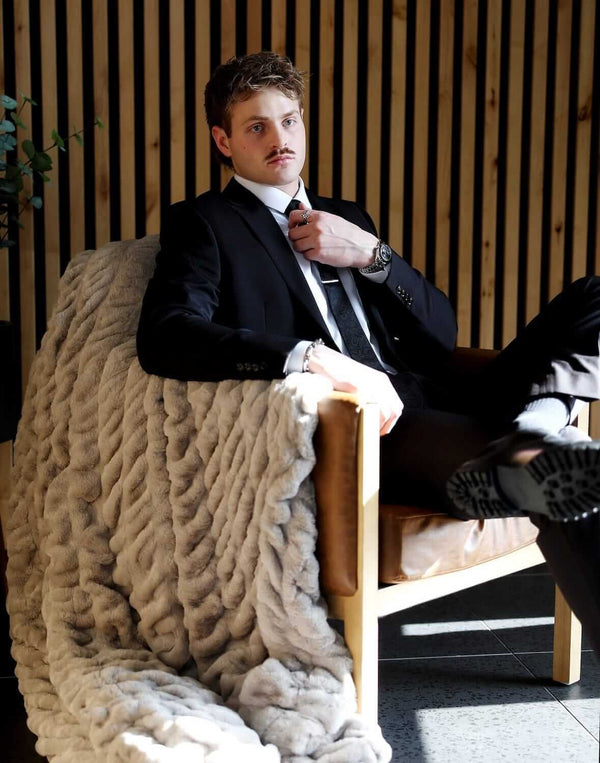A man in a suit sitting on a chair, draped with a Biggest Hugs Tranquil Dove Blanket, showcasing its luxurious texture.