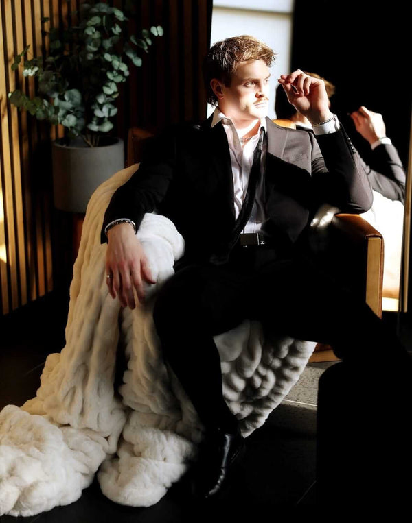 A stylish man in a black suit lounging on a chair covered with the Biggest Hugs Cream weighted blanket.