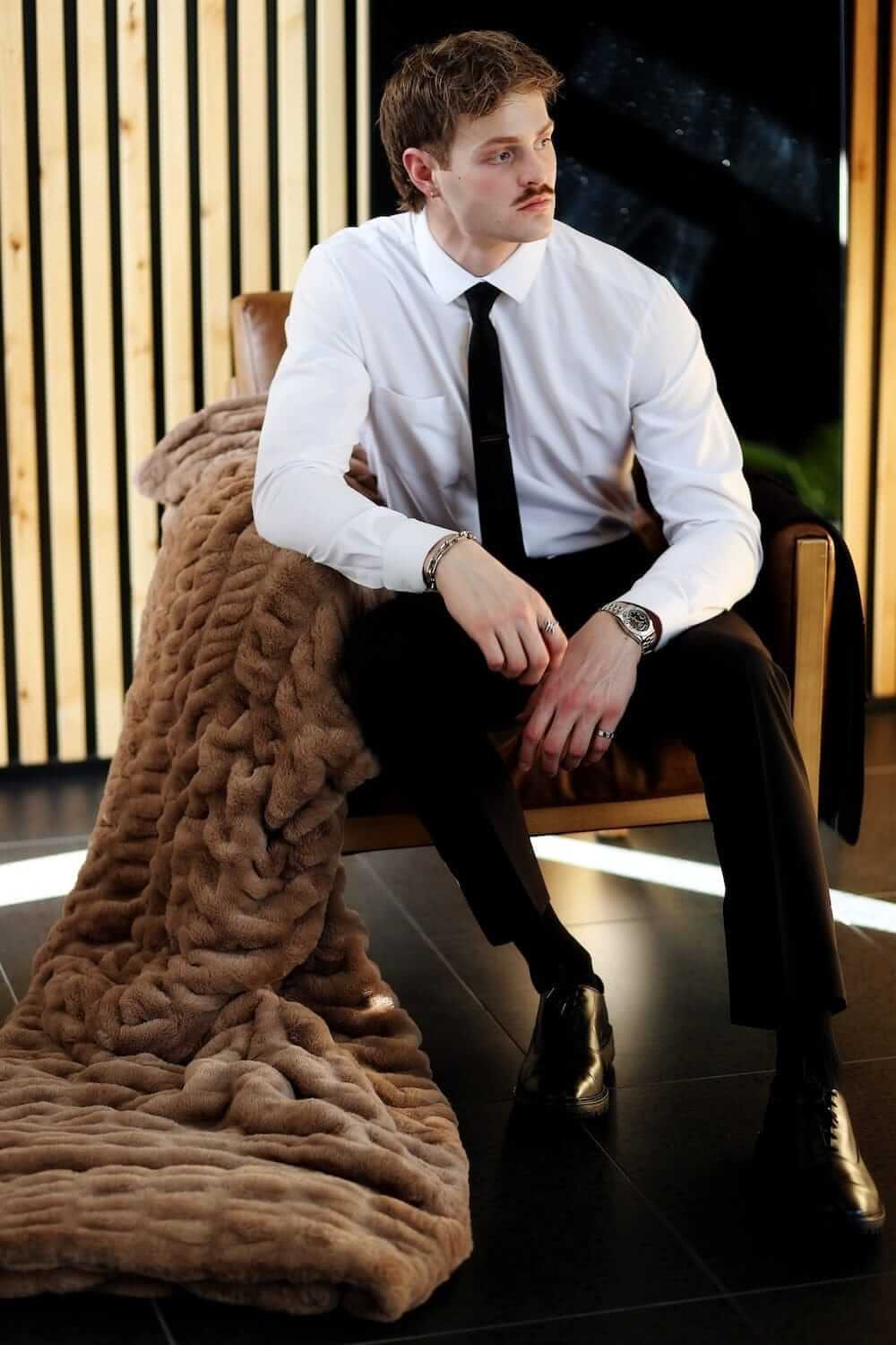 Stylish man seated with Biggest Hugs Milk Chocolate weighted blanket draped over a chair, showcasing its soft texture and warm color.