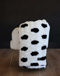 White Minky Hugs Polka Dot Marshmallow plush blanket with black spots draped over a cream chair.